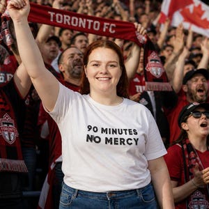 Puede incluir: Una camiseta blanca con el texto "90 MINUTES NO MERCY" en negro, usada por una persona en una multitud de aficionados vitoreando. La multitud sostiene bufandas y pancartas rojas con "TORONTO FC" impreso.