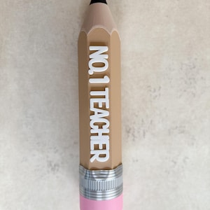 May include: A pencil-shaped object with a tan body, a black tip, and a pink eraser at the base. A silver-coloured metal band separates the eraser from the body. The white text NO. 1 TEACHER is embossed vertically along the centre of the tan pencil.