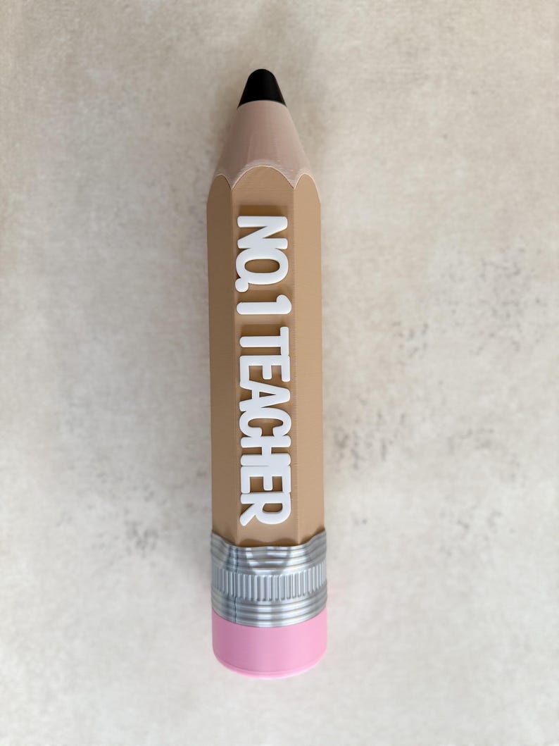 May include: A pencil-shaped object with a tan body, a black tip, and a pink eraser at the base. A silver-coloured metal band separates the eraser from the body. The white text NO. 1 TEACHER is embossed vertically along the centre of the tan pencil.