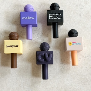 May include: Five small microphone-shaped charms in various colors. One is purple with 'mellow', one black with 'ECC', one yellow with 'honeycuff', one dark blue with 'CV', and one light pink with 'TINY JOY KITS' and a sun icon.