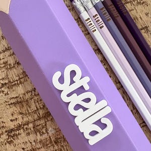 May include: A large, light purple pencil-shaped container with the name Stella in white lettering, accompanied by four standard-sized pencils in shades of white, light purple, dark purple, and deep plum, each personalised with the name Stella.