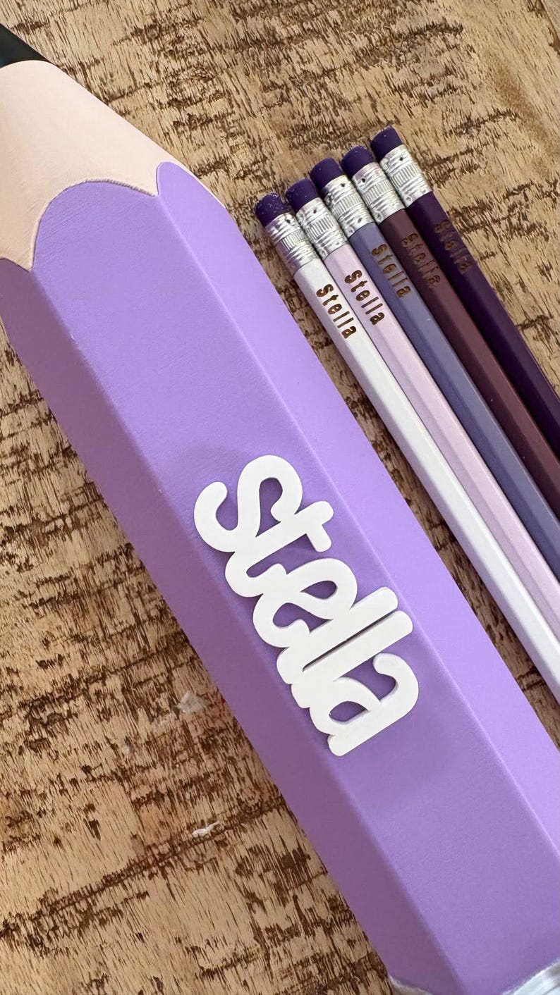 May include: A large, light purple pencil-shaped container with the name Stella in white lettering, accompanied by four standard-sized pencils in shades of white, light purple, dark purple, and deep plum, each personalised with the name Stella.