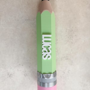 May include: A light green pencil-shaped object featuring a black tip, a tan wooden-coloured section, and a pink eraser at the base with a silver-coloured metal band. The name Lucas is embossed in white capital letters vertically along the green body.