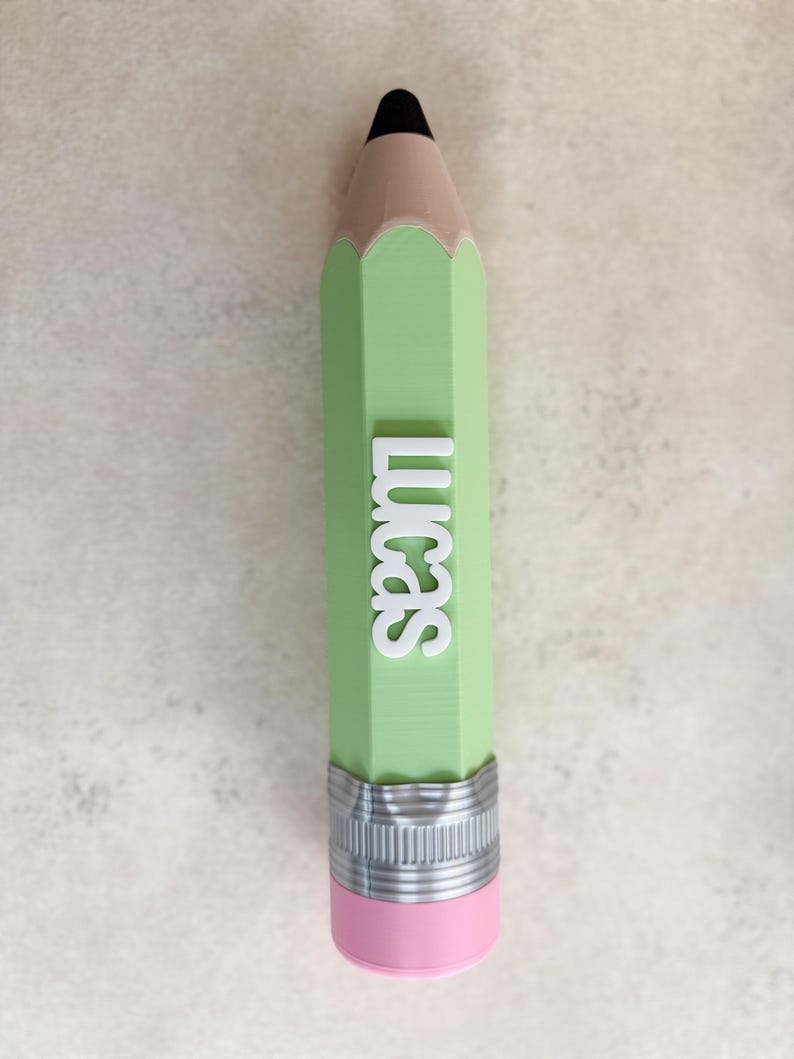 May include: A light green pencil-shaped object featuring a black tip, a tan wooden-coloured section, and a pink eraser at the base with a silver-coloured metal band. The name Lucas is embossed in white capital letters vertically along the green body.