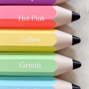 May include: A vertical stack of seven large pencil-shaped objects in light brown, purple, hot pink, yellow, green, baby blue, and denim blue. Each object features a tan wooden tip and a black lead point, with the colour name printed in white text.