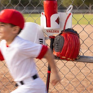 May include: A baseball setup with a red water bottle, glove, helmet, and bat resting on a chain-link fence. The white holder has the word "VALDES" printed on it. A person in a baseball uniform is running in the foreground.