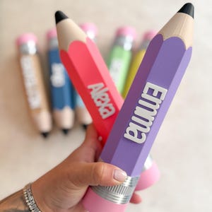 May include: A collection of pencil-shaped containers in various colours including purple, red, blue, green, and tan. Each pencil features a black tip, a silver-coloured band, and a pink eraser end. The purple pencil displays the name Emma, and the red pencil displays the name Alexa in raised white lettering.