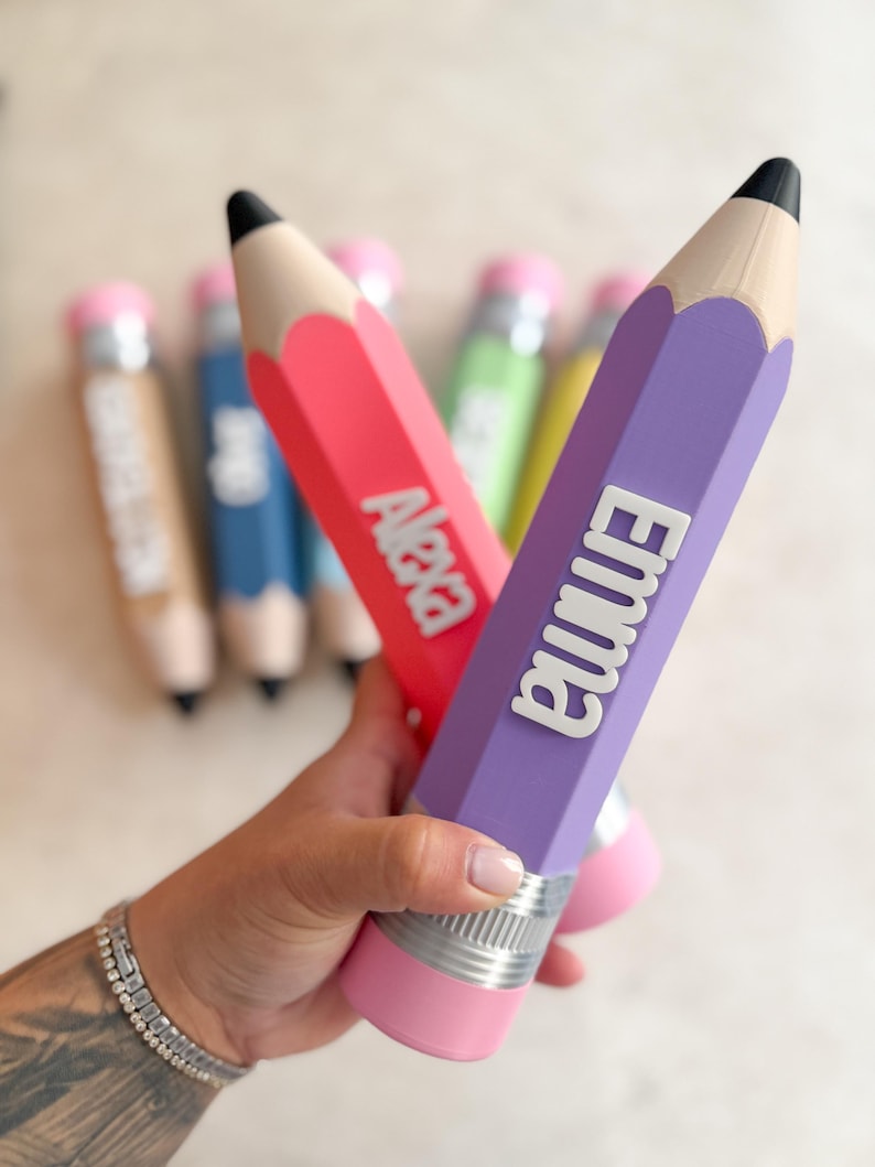May include: A collection of pencil-shaped containers in various colours including purple, red, blue, green, and tan. Each pencil features a black tip, a silver-coloured band, and a pink eraser end. The purple pencil displays the name Emma, and the red pencil displays the name Alexa in raised white lettering.