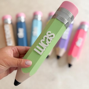 May include: A light green pencil-shaped container with the name LUCAS in raised white letters. It features a pink eraser top, a silver-coloured metal band, and a tan wooden-coloured base with a black tip. Other similar containers are in the background.