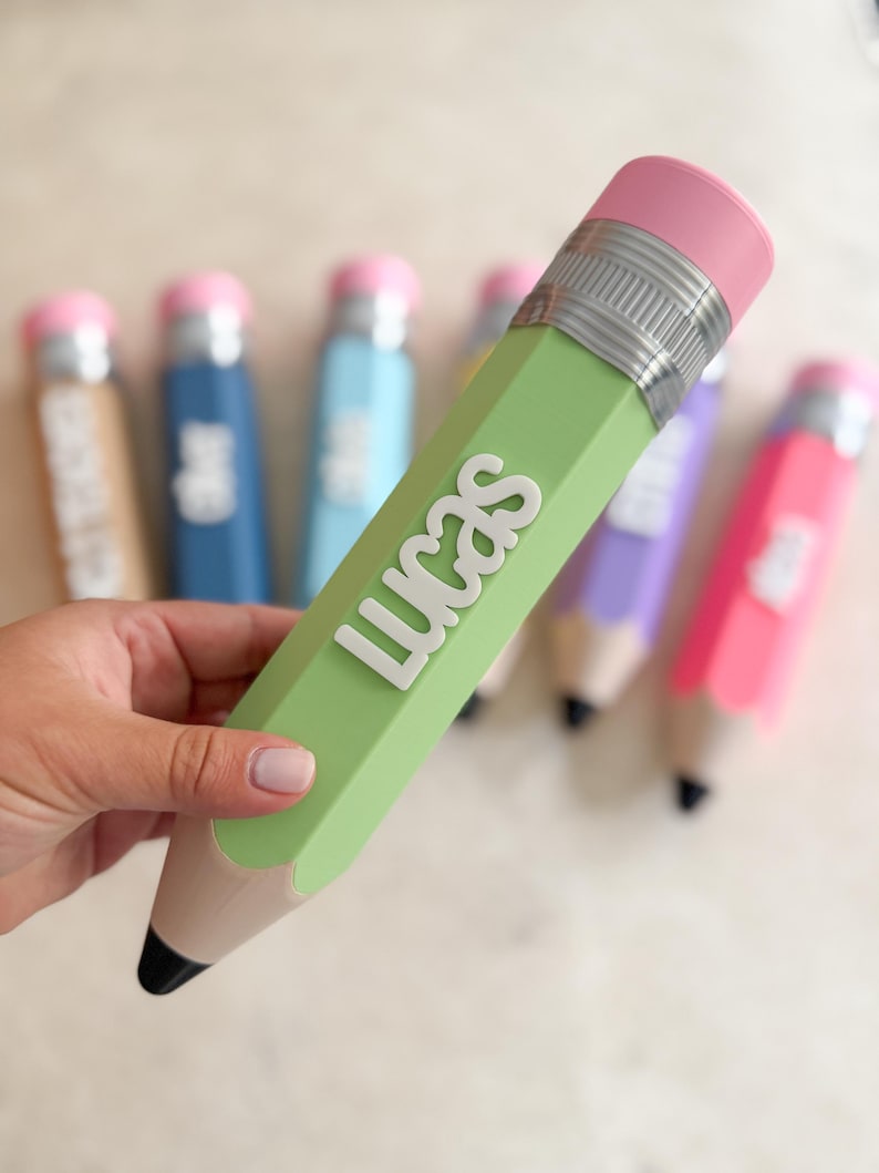 May include: A light green pencil-shaped container with the name LUCAS in raised white letters. It features a pink eraser top, a silver-coloured metal band, and a tan wooden-coloured base with a black tip. Other similar containers are in the background.