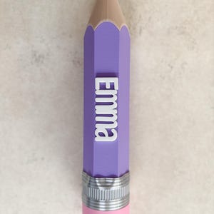 May include: A purple pencil-shaped object featuring a black tip, a tan wooden-coloured section, and a silver-coloured metal ferrule with a pink eraser at the base. The name Emma is displayed in raised white lettering along the purple body.