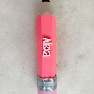 May include: A pink pencil-shaped object featuring a black tip, a silver-coloured ferrule, and a light pink eraser at the base. The name Alexa is written in white, raised, block-style lettering vertically along the centre of the pink pencil body.