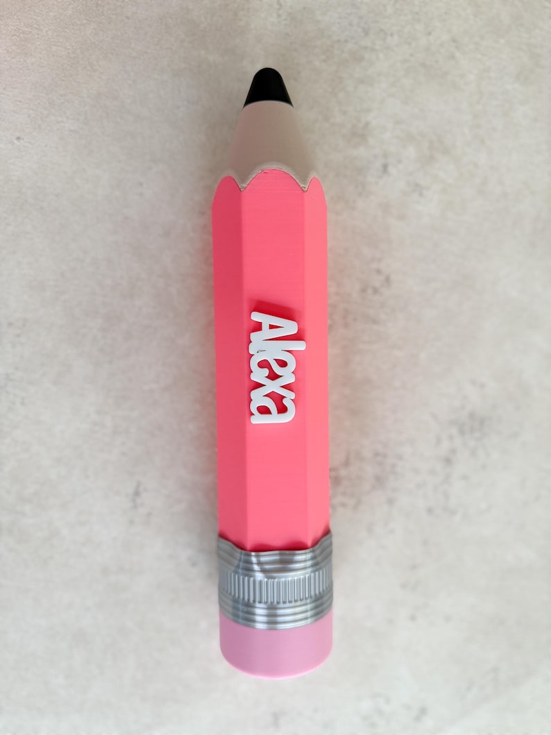 May include: A pink pencil-shaped object featuring a black tip, a silver-coloured ferrule, and a light pink eraser at the base. The name Alexa is written in white, raised, block-style lettering vertically along the centre of the pink pencil body.
