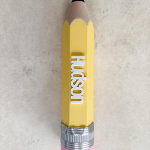 May include: A yellow pencil-shaped object with a black tip, a tan wooden-coloured section, and a silver metal ferrule with a pink eraser at the base. The name Hudson is embossed in white, raised capital letters vertically along the yellow body.