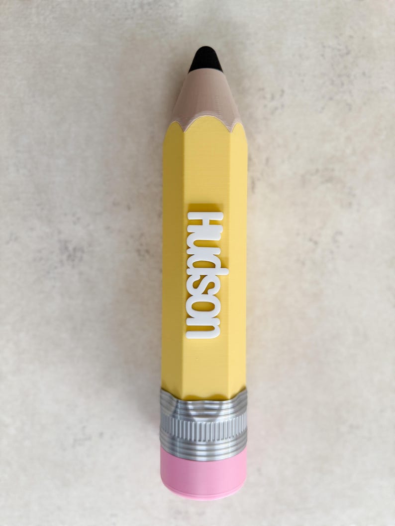 May include: A yellow pencil-shaped object with a black tip, a tan wooden-coloured section, and a silver metal ferrule with a pink eraser at the base. The name Hudson is embossed in white, raised capital letters vertically along the yellow body.