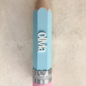 May include: A light blue pencil-shaped object featuring a black tip, a tan wooden-coloured section, and a silver-coloured metal ferrule above a pink eraser. The name Olivia is embossed in white, raised, sans-serif capital letters on the side.