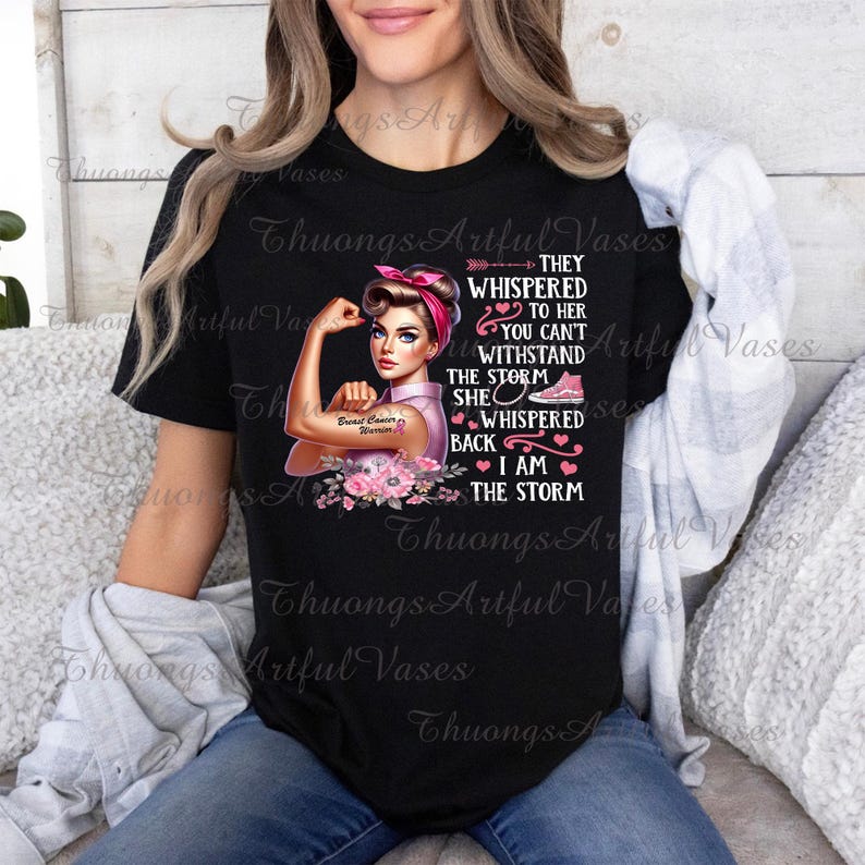 Puede incluir: Camiseta negra con un gr&aacute;fico rosa y blanco de una mujer en una pose de brazo fuerte. El gr&aacute;fico incluye el texto "Breast Cancer Warrior" y "They whispered to her you can't withstand the storm. She whispered back I am the storm".