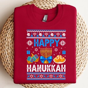Może przedstawiać: Czerwona bluza z okazji Chanuki z uroczystym wzorem. Wz&oacute;r zawiera słowa "HAPPY HANUKKAH" w kolorze niebieskim i białym, wraz z menorami, bączkami i innymi symbolami świątecznymi. Bluza wykonana jest z miękkiego materiału.