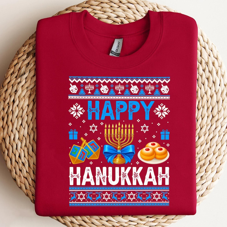 Może przedstawiać: Czerwona bluza z okazji Chanuki z uroczystym wzorem. Wz&oacute;r zawiera słowa "HAPPY HANUKKAH" w kolorze niebieskim i białym, wraz z menorami, bączkami i innymi symbolami świątecznymi. Bluza wykonana jest z miękkiego materiału.
