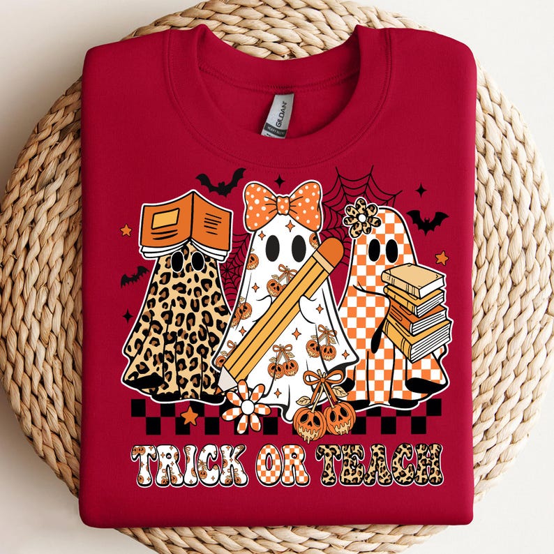 May include: A red sweatshirt featuring three ghost characters with Halloween-themed designs. One ghost has a leopard print, another holds a pencil, and the third carries books. The text "TRICK OR TEACH" is displayed at the bottom.