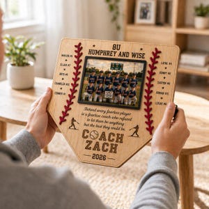 May include: Wooden baseball home plate-shaped photo frame. The frame features a team photo, the text "COACH ZACH - 2026", and player names. Red stitching and baseball silhouettes decorate the edges. The frame is held by a person.