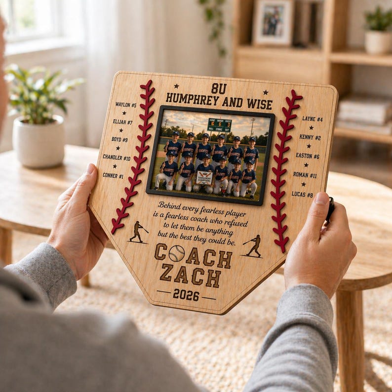 May include: Wooden baseball home plate-shaped photo frame. The frame features a team photo, the text "COACH ZACH - 2026", and player names. Red stitching and baseball silhouettes decorate the edges. The frame is held by a person.
