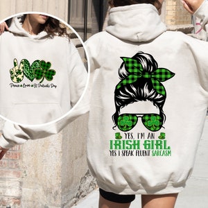 May include: A white hoodie with a green shamrock design and the text "Peace & Love & St Patrick's Day" on the front. The back of the hoodie features a cartoon illustration of a woman with a green plaid bun, sunglasses with shamrocks, and the text "YES, I'M AN IRISH GIRL YES I SPEAK FLUENT SARCASM".