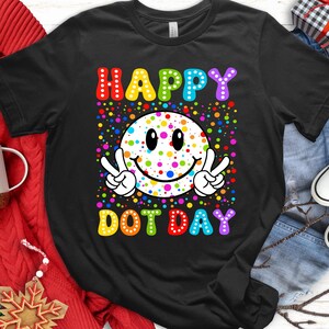 May include: Black t-shirt with a colorful graphic celebrating "Happy Dot Day." The design features a smiley face with peace signs, surrounded by dots, and the words "HAPPY DOT DAY" in rainbow colors.