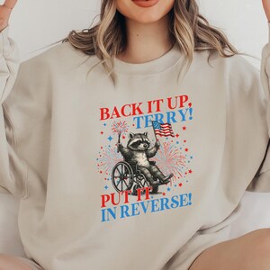 May include: Beige sweatshirt with a graphic of a raccoon in a wheelchair holding an American flag. The text "BACK IT UP, TERRY! PUT IT IN REVERSE!" is printed above and below the raccoon illustration, with red, white, and blue accents.