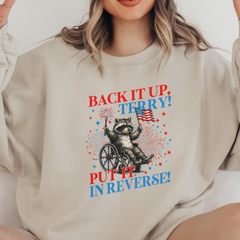 May include: Beige sweatshirt with a graphic of a raccoon in a wheelchair holding an American flag. The text "BACK IT UP, TERRY! PUT IT IN REVERSE!" is printed above and below the raccoon illustration, with red, white, and blue accents.