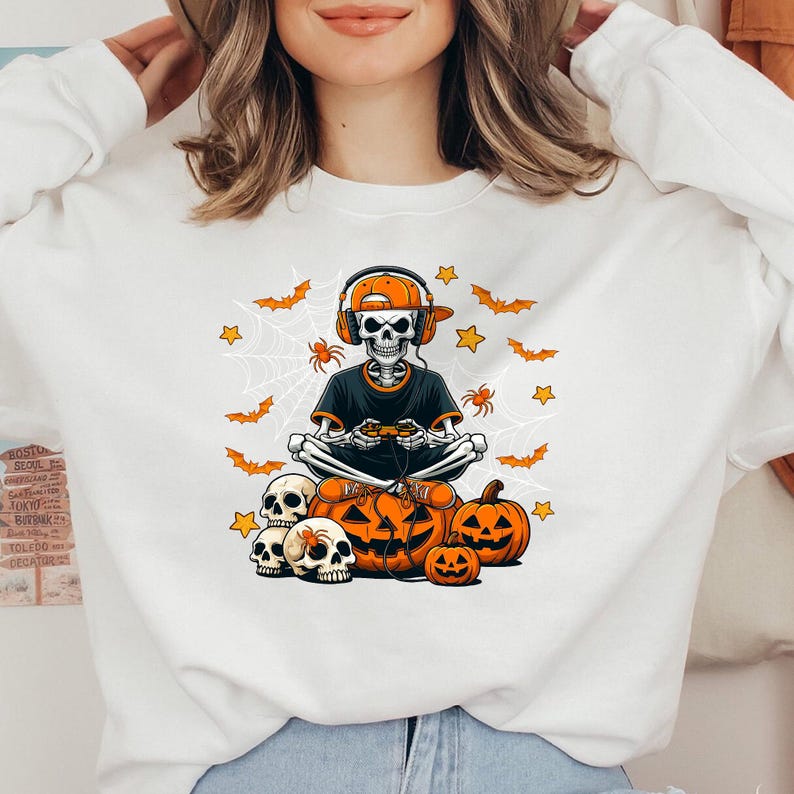 May include: White sweatshirt with a Halloween graphic. The design shows a skeleton wearing headphones and holding a game controller, with pumpkins, bats, spiders, and stars. The color scheme is orange, black, and white.