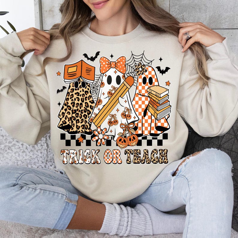 May include: Beige sweatshirt with a Halloween-themed graphic. The design features three ghost characters, one in leopard print, one with a bow, and one in orange checkered pattern. The phrase "TRICK OR TEACH" is printed below the ghosts.
