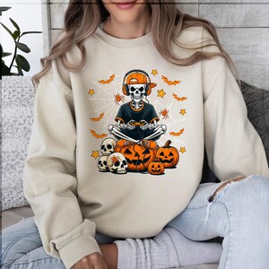 May include: Beige sweatshirt with a Halloween design. The graphic shows a skeleton wearing headphones and playing a game, with pumpkins, bats, spiders, and stars. A casual, comfortable style of sweatshirt.