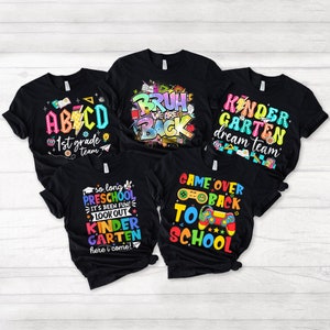 May include: Five black t-shirts with colourful text and graphics. The shirts feature phrases like "Kindergarten Dream Team", "Game Over Back to School", "So Long Preschool", "Bruh We Are Back", and "ABC 1st Grade Team".