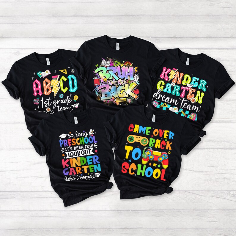 May include: Five black t-shirts with colourful text and graphics. The shirts feature phrases like "Kindergarten Dream Team", "Game Over Back to School", "So Long Preschool", "Bruh We Are Back", and "ABC 1st Grade Team".