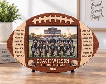May include: A football-shaped wooden picture frame featuring a central photo of a team in black uniforms. The frame displays the text "COACH WILSON TIGERS FOOTBALL 2027" and lists player names with jersey numbers on the left and right sides.