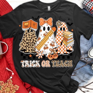 May include: Black t-shirt with a Halloween-themed design. Features three ghost figures, one with a leopard print, one with a bow and pencil, and one with a checkered pattern. The text "TRICK OR TEACH" is displayed in a decorative font.