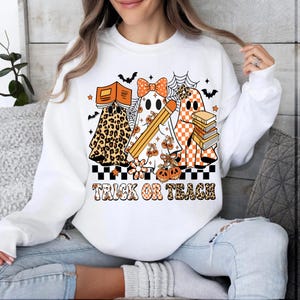 May include: White sweatshirt with a Halloween-themed graphic. The design features three ghost characters with books, pencils, and pumpkins. The text "TRICK OR TEACH" is printed in orange and black.