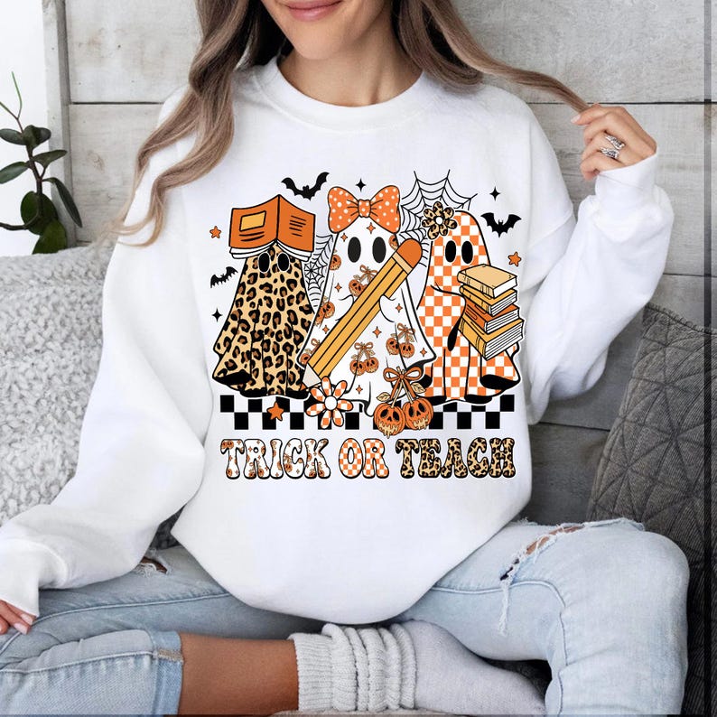 May include: White sweatshirt with a Halloween-themed graphic. The design features three ghost characters with books, pencils, and pumpkins. The text "TRICK OR TEACH" is printed in orange and black.