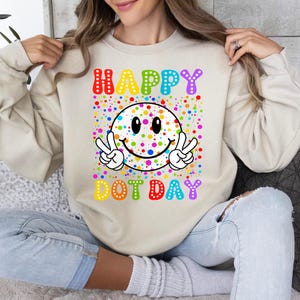 May include: Beige sweatshirt with a colorful graphic. The graphic features a smiley face with peace signs, surrounded by colorful dots. The words "HAPPY DOT DAY" are written above and below the smiley face in rainbow colors.