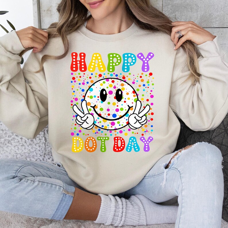 May include: Beige sweatshirt with a colorful graphic. The graphic features a smiley face with peace signs, surrounded by colorful dots. The words "HAPPY DOT DAY" are written above and below the smiley face in rainbow colors.