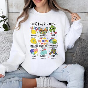 May include: White sweatshirt with a colorful graphic design that says "God says I am..." and lists positive attributes with bible verses. The graphic includes a hand, butterfly, puzzle piece, lightbulb, brain, heart, boxing glove, and a ribbon.