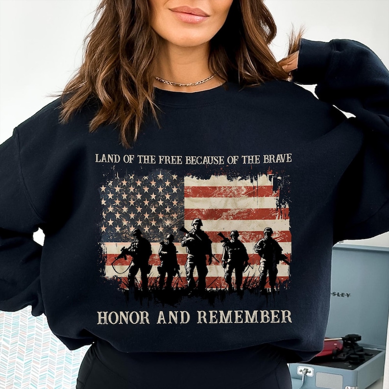 May include: Black sweatshirt featuring an American flag design with soldiers and the phrases "LAND OF THE FREE BECAUSE OF THE BRAVE" and "HONOR AND REMEMBER" in white. The flag has red and white stripes and a field of stars.