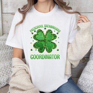 May include: White t-shirt with a green four-leaf clover design and the text "PRESCHOOL SHENANIGANS COORDINATOR".