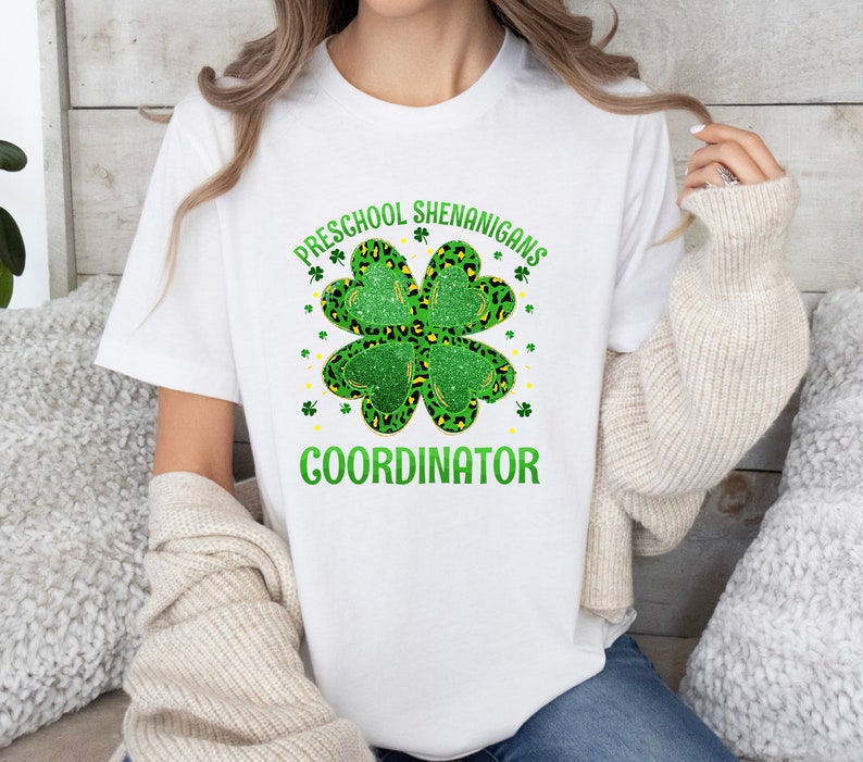 May include: White t-shirt with a green four-leaf clover design and the text "PRESCHOOL SHENANIGANS COORDINATOR".