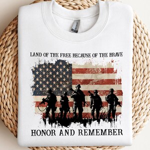 May include: White sweatshirt featuring a distressed American flag design with the text "Land of the Free Because of the Brave" and "Honor and Remember." The graphic includes silhouettes of soldiers holding rifles.