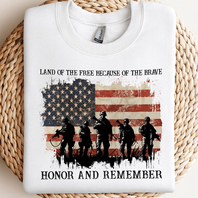 May include: White sweatshirt featuring a distressed American flag design with the text "Land of the Free Because of the Brave" and "Honor and Remember." The graphic includes silhouettes of soldiers holding rifles.