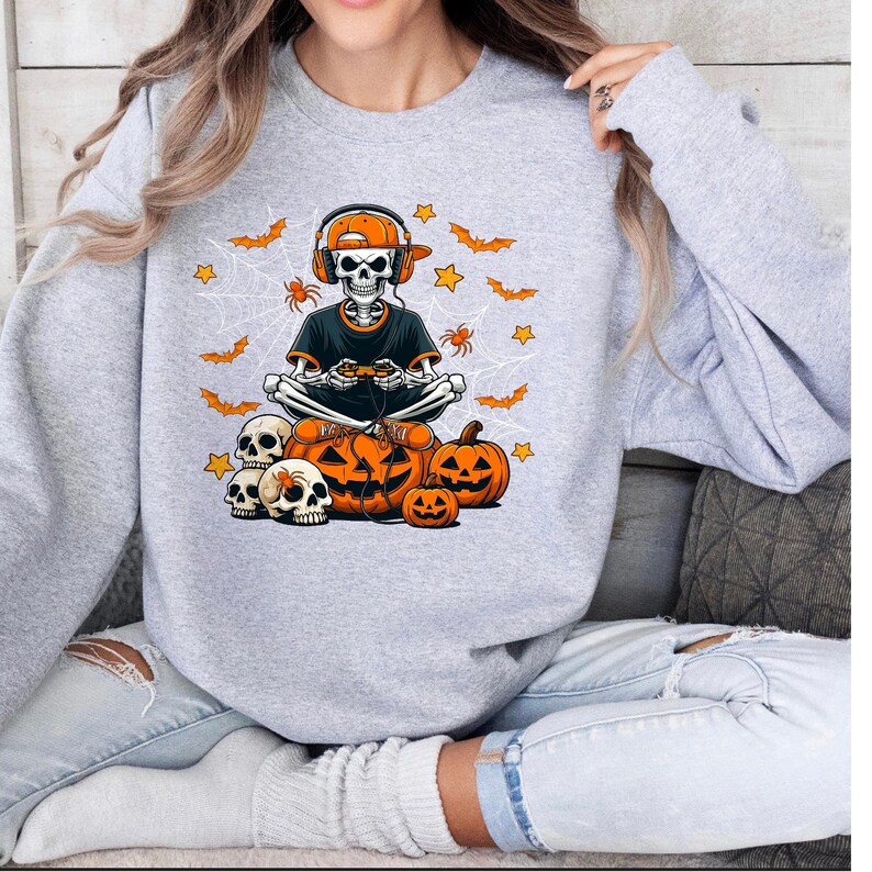May include: A light grey sweatshirt with a Halloween design. The graphic shows a skeleton wearing headphones and a cap, holding a games controller, with pumpkins, bats, spiders, and stars.