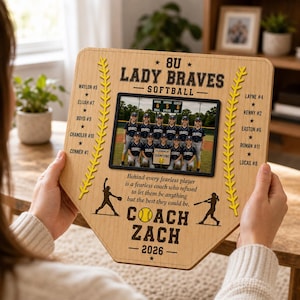 May include: A wooden baseball home plate-shaped plaque featuring a team photo and text. The plaque includes the text "8U Lady Braves Softball" and "Coach Zach - 2026". The team photo is in a black frame. Yellow stitching and star accents are also present.