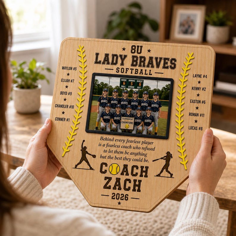 May include: A wooden baseball home plate-shaped plaque featuring a team photo and text. The plaque includes the text "8U Lady Braves Softball" and "Coach Zach - 2026". The team photo is in a black frame. Yellow stitching and star accents are also present.